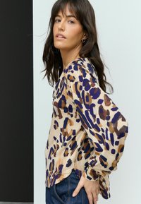 Blouse en imprimé léopard beige, avec des motifs violets et marron, à manches longues et coupe décontractée. Tissu lisse, ourlet arrondi.