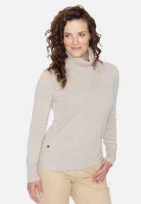 Beige Rollkragenpullover mit strukturiertem Strickmuster und gerippten Bündchen. Auffälliger seitlicher Akzentknopf fügt ein Detail hinzu. Das Model trägt helle Hosen.