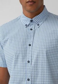 Chemise à manches courtes de couleur bleu clair avec un motif géométrique. Elle présente un col boutonné et des boutons bleu foncé. Le motif est constitué de petits carrés.