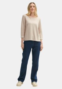 Hellbeige Langarmoberteil mit strukturierter Oberfläche, kombiniert mit dunkelblauen geraden Jeans und beigen Sneakers. Schlichtes, lässiges Design.