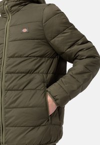 Oliwkowa ocieplana kurtka z zamkiem błyskawicznym z przodu, poziomym pikowaniem i boczną kieszenią; wykonana z gładkiej, fakturowanej tkaniny.