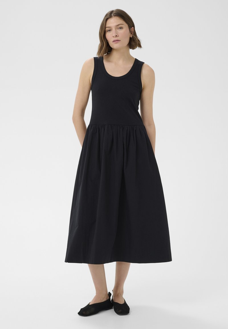 Femme debout, mains dans les poches, portant une robe midi noire sans manches et des ballerines noires sur un fond gris uni.