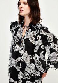 Schwarze transparente Bluse mit weißen Paisley- und Blumenmustern, mit gerafftem Ausschnitt, Bindeverschluss und voluminösen Ärmeln.