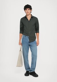 Camisa de botones verde oscuro, jeans azul claro, zapatillas negras, sosteniendo una bolsa de mano beige. Ajuste casual con mangas largas y una textura suave.