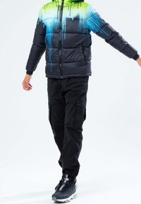 Zwarte pufferjas met een gradient ontwerp van groen naar blauw drip, gecombineerd met zwarte cargobroeken en donkere sneakers met witte accenten.