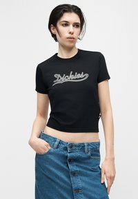 Νεαρό άτομο με κοντά σκούρα μαλλιά φορεί ένα μαύρο κοντό T-shirt με το λογότυπο "Dickies" και μια μπλε τζιν φούστα με κουμπιά, ποζάροντας μπροστά σε λευκό φόντο.