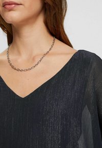 Vestito nero con scollo a V realizzato in un tessuto testurizzato e luccicante. Presenta una vestibilità ampia e una collana in argento con anelli ovali attorno al collo.