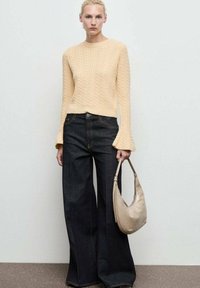 Donna con maglione beige a trecce, jeans neri a gamba larga, che tiene una borsa a tracolla beige, in piedi di fronte a un muro bianco semplice.