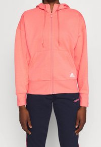Felpa rosa zip-up con tasca frontale, cappuccio con cordino e polsini a costine. Presenta il logo Adidas in bianco in basso a sinistra.