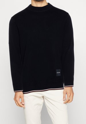 Mand iført en sort langærmet sweater med ribbet tekstur og hvide bukser, sweateren har hvide og røde kantbånd ved ærmer og kant.