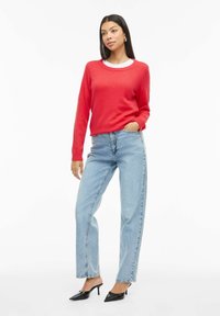 Femme debout, main dans la poche, portant un pull rouge, un jean bleu clair et des escarpins noirs à bout pointu sur fond blanc.