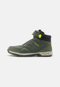Chaussure haute synthétique verte avec col noir, accents jaunes et semelle tachetée. Présente un design à lacets et un détail de logo sur le côté.