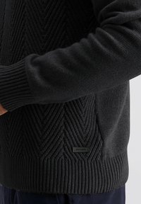 Schwarzer Pullover mit einem strukturierten Zickzackmuster, gerippten Bündchen und Saum. Verfügt über ein kleines Markenetikett an der Seite. Weicher Stoff, normale Passform.