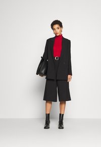 Svart skreddersydd blazer over en rød ribbestrikket turtleneck, kombinert med svarte vide shorts og svarte ankelboots. Tilbehøret inkluderer en stor håndveske.