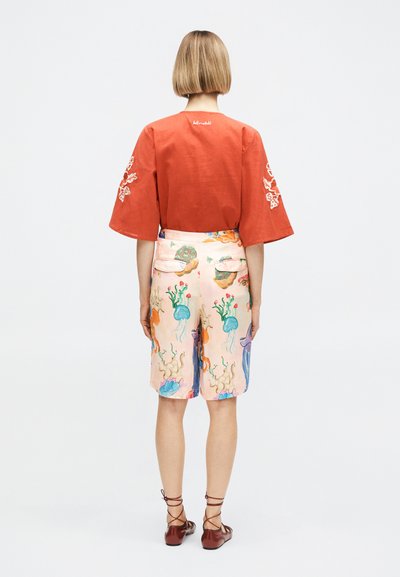 Personne aux cheveux blonds courts, vue de dos, portant un haut orange rouille avec une broderie blanche sur les manches et un short pastel à imprimés marins colorés.