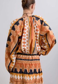 Vestido fluido y con estampado que presenta colores naranja, negro y blanco. Manga largas y sueltas, cintura ceñida y falda en capas con diseños distintivos.