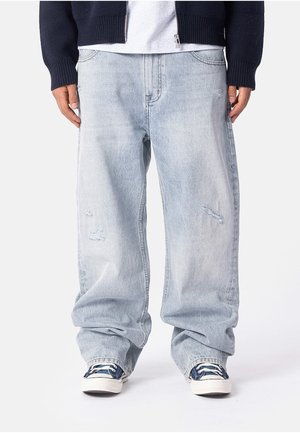 Person, die hellblaue, gewaschene, locker sitzende Jeans mit leichten Gebrauchsspuren, marineblaue und weiße Sneakers sowie eine dunkelblaue Jacke mit einem weißen Hemd trägt.