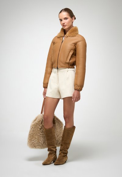 Giubbotto bomber in pelle marrone con colletto in pelliccia, pantaloni corti crema, stivali marroni in suede al ginocchio e una grande borsa in peluche beige.