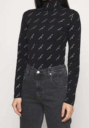 Femme portant un top col roulé noir à manches longues avec de petits logos blancs répétés « Calvin Klein Jeans » et un jean gris foncé taille haute.