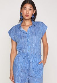 Camicia a maniche corte a righe blu, con chiusura frontale a bottoni e vita elastica. Presenta una tasca e un colletto; il tessuto appare leggero e testurizzato.
