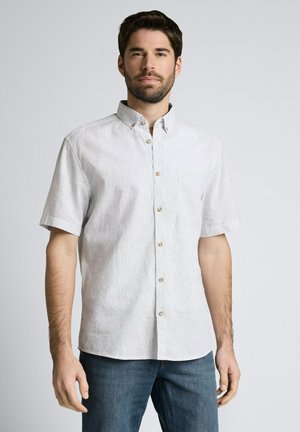 Mann mit dunklem Haar und Bart, der ein kurzärmeliges, helles Hemd mit Knöpfen und blaue Jeans trägt, steht vor einem schlichten Hintergrund.