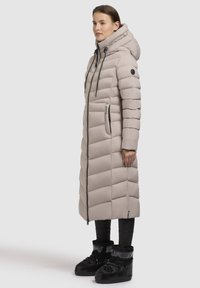 Long beige puffer coat with vertical seams, adjustable hood, front zipper, side pockets, worn over black insulated boots.