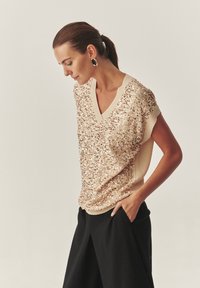 Bronzas krāsas topiņš ar spīdīgām pērlītēm, V veida izgriezumu un īsām piedurknēm, kombinēts ar melnām augsta vidukļa biksēm. Gluda tekstūra bez redzamām rakstu formām.