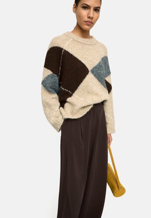 Kobieta w beżowym swetrze z dużymi brązowo-niebieskimi wzorami diamentowymi, szerokich ciemnobrązowych spodniach, trzymająca małą żółtą torebkę.