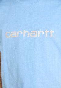 Light blue cotton t-shirt featuring a prominent white "carhartt" logo in a bold, modern font on the front. Short sleeves, crew neck.