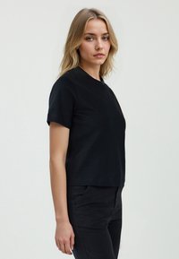 Blondynka w czarnym t-shircie i czarnych spodniach stoi na jasnym tle, patrząc w stronę kamery z neutralnym wyrazem twarzy.