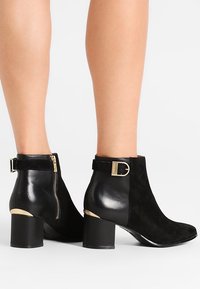 Bottes courtes en cuir noir et daim avec boucle dorée, zip et talons carrés portées sur des jambes nues sur fond blanc.