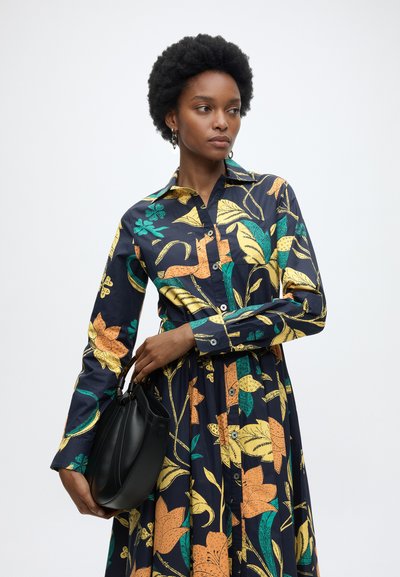 Femme aux cheveux courts et bouclés portant une robe noire à manches longues avec des motifs floraux jaunes et verts, tenant un sac à main noir.