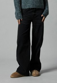 Svarta byxor med vida ben i denim, tillsammans med en figursydd tröja i teal och bruna tofflor. Tröjan har långa ärmar med ribbade manschetter.