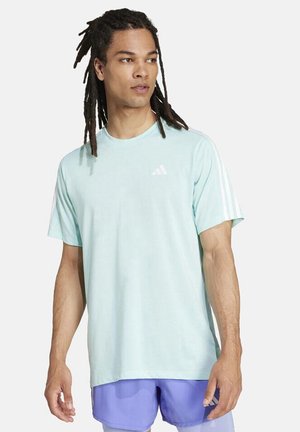 Man met lange dreadlocks, een lichtblauw Adidas T-shirt en paarse sportshorts, die iets naar links kijkt tegen een witte achtergrond.