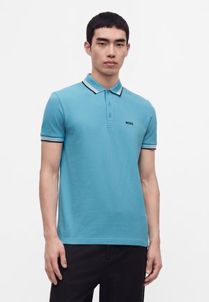 Ung mand med kort sort hår iført en lyseblå poloshirt med sort og hvid kant og sorte bukser, stående mod en ensfarvet baggrund.