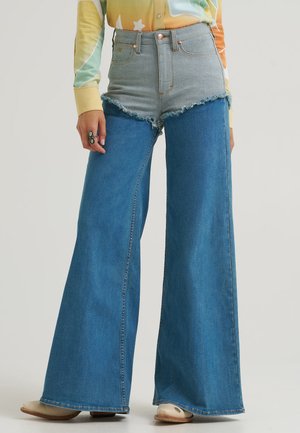 Personne portant un jean à jambes larges avec un short taille haute bleu clair superposé sur un pantalon large bleu foncé et des chaussures beiges.