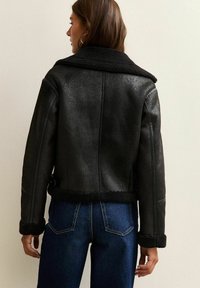 Giacca in shearling nera con colletto, maniche con polsini e vita con cintura. Finitura in pelle texture, design corto e cuciture a vista.