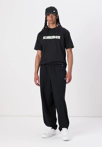 Čierne tričko s nápisom "NICE CONDITIONS GEAR", kombinované s voľnými čiernymi nohavicami a bielymi teniskami. Obsahuje čiernu čiapku.