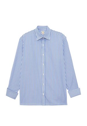 Personne avec de longs cheveux tressés portant une chemise à manches longues rayée verticalement bleue et blanche et un pantalon marron clair, vue de dos.