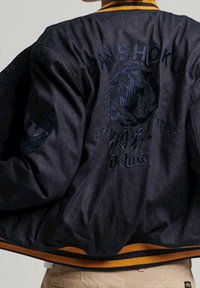 Veste bomber noire avec des détails brodés d'un tigre et de texte. Col et poignets côtelés jaunes. Tissu lisse avec un léger éclat.