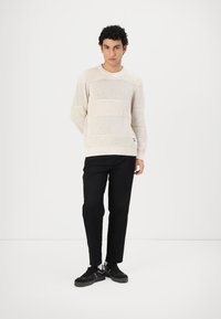 Suéter de punto beige con rayas horizontales, cuello redondo y dobladillo acanalado, combinado con pantalones negros y zapatillas negras.