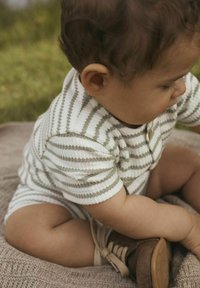 Dryžuotas bodysuit iš balto ir alyvuogių žalumo audinio, su trumpomis rankovėmis ir apvali iškirpte, derinamas su rudos odos bateliais.