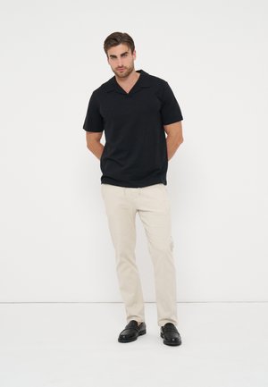 Man draagt een zwart overhemd met korte mouwen en kraag, beige broek en zwarte loafers, staat met handen achter de rug tegen een witte achtergrond.