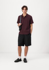 Korte mouwen, bordeaux geruit overhemd, zwarte shorts, zwarte instappers, witte sokken. Model houdt een kleine zwarte schoudertas vast.