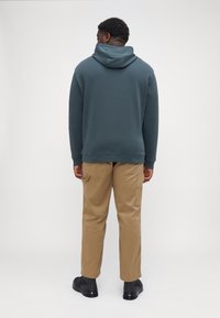Sudadera con capucha de algodón verde oscuro con un corte relajado, combinada con pantalones cargo beiges y calzado negro, vista desde la parte posterior.