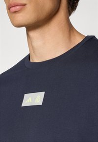 Camiseta de algodón azul marino con cuello redondo, que presenta un logo impreso en gris claro y amarillo en la parte frontal. Textura suave, corte casual.
