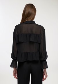 Blusa nera trasparente con balze arricciate, maniche lunghe con polsini in elastico e colletto. Il tessuto ha una consistenza leggera e fluida.