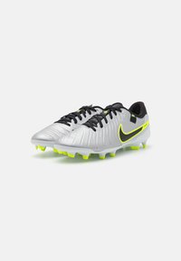 Bottes de football Nike argentées avec des accents noirs, tige texturée, détails jaune-vert et crampons moulés pour une traction sur les surfaces en herbe.