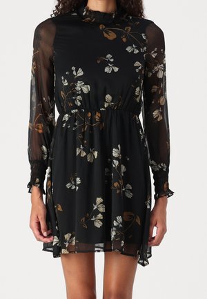 Sort blomsterkjole med gennemsigtige lange ærmer, samlet talje og flæsekantede manchetter, båret af en person med krøllet hår.