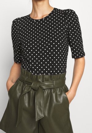 Sort top med hvide prikker, korte ærmer, rund halsudskæring og rynker ved skuldrene, kombineret med olivengrønne højtaljede shorts i kunstlæder med bælte i taljen.